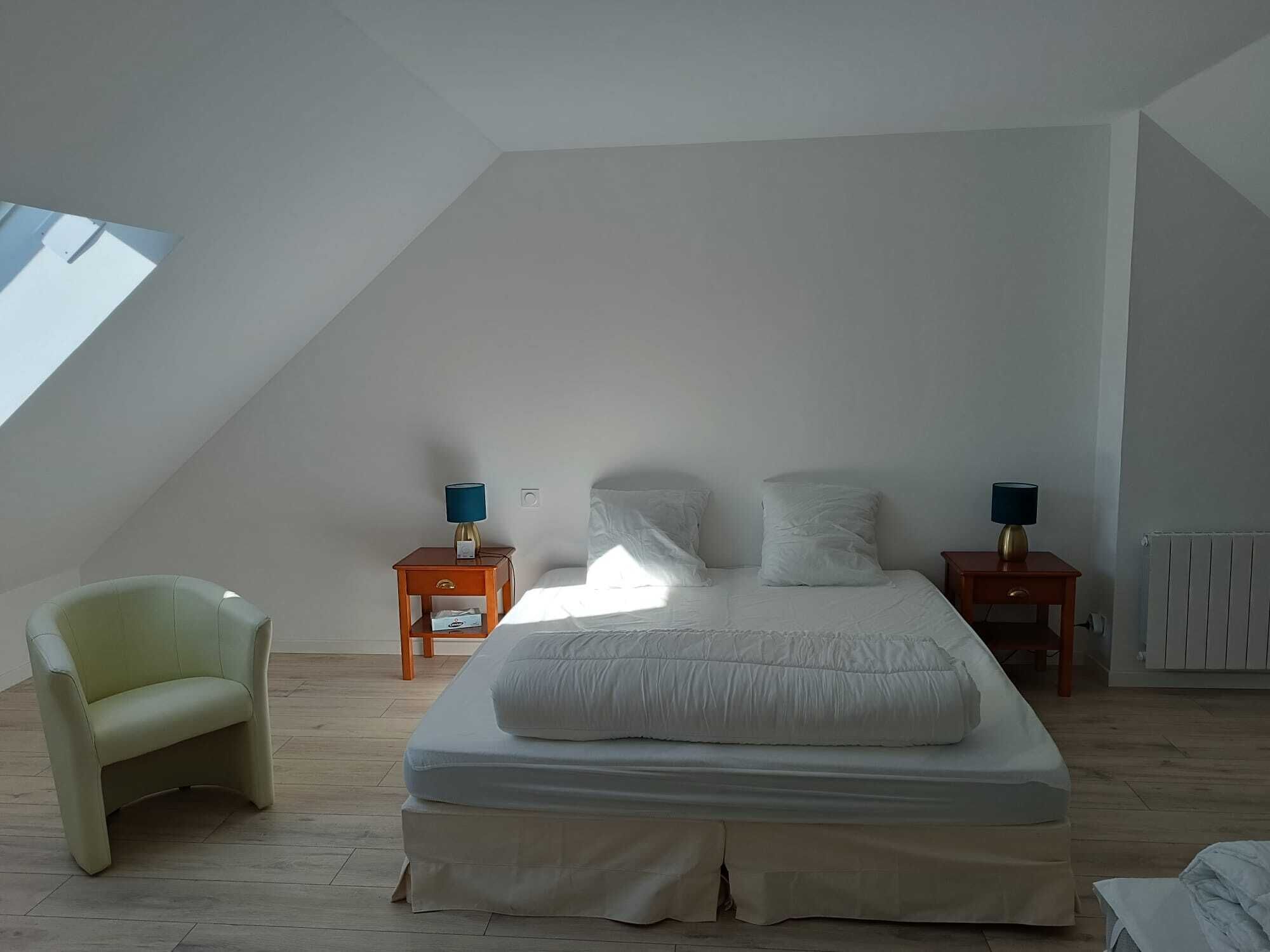 Un dormitorio con techo inclinado, una cama con ropa de cama blanca, dos mesitas de noche con lámparas y una sola silla.