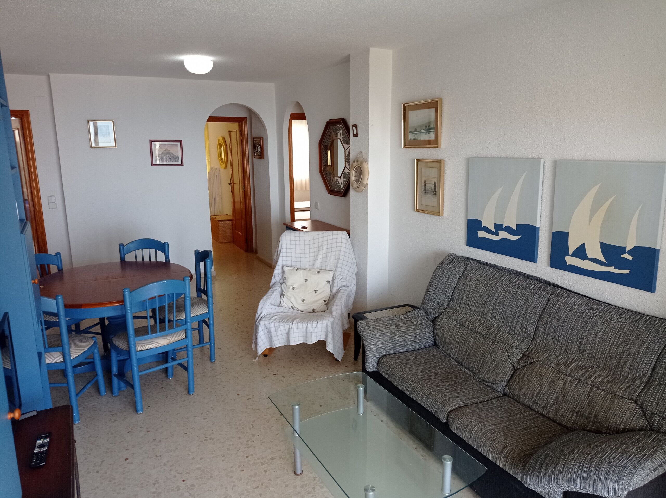 A living room with a sofa, a glass coffee table, a dining table with chairs, and a hallway leading to another room.