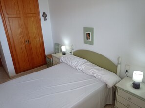 A bedroom with a bed, bedside tables, a nightstand, a wooden wardrobe, a wall-mounted cross, and a framed picture. - Mar Azul  6º, Solo Familias! (Playa Xeraco)
