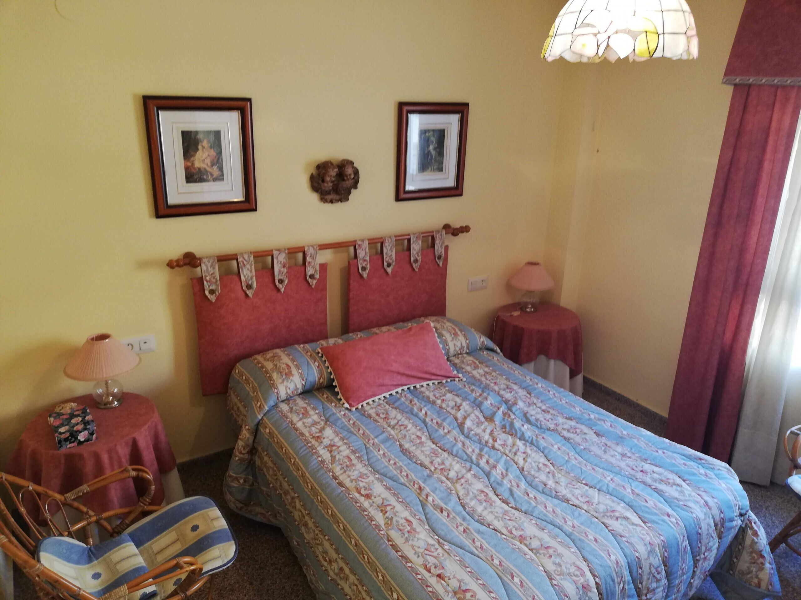 Une chambre à coucher avec un lit, deux tableaux encadrés au mur, une table de chevet avec une lampe, une chaise et une petite table avec une lampe.