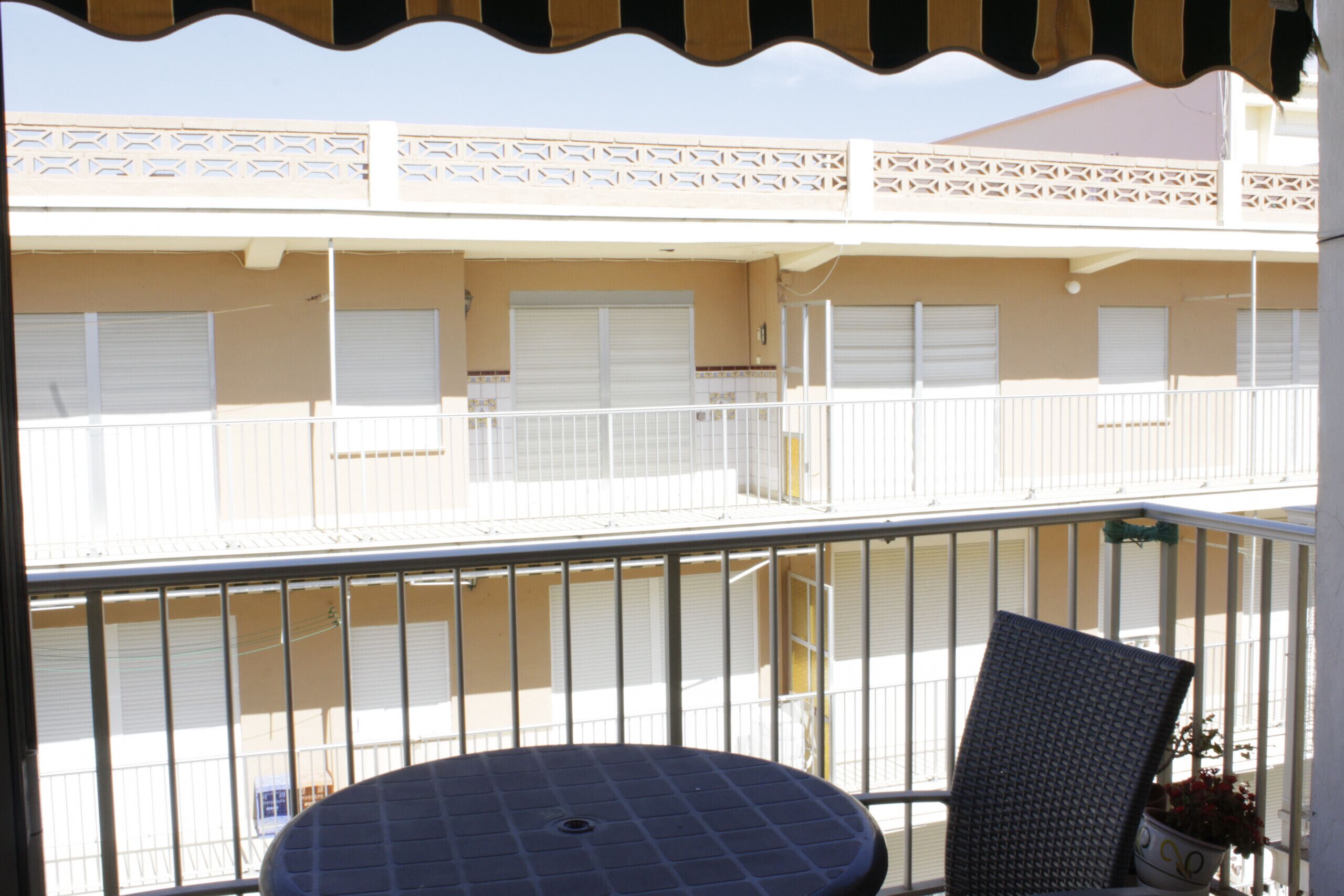 A balcony with a round table and a chair, overlooking a building with closed shutters and a decorative railing.