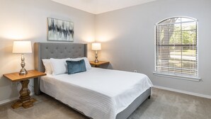 A bedroom with a large bed, two bedside tables with lamps, a wall-mounted artwork, and a window with blinds.