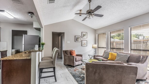 A modern kitchen with a granite countertop, a dining area with a chair and a sofa, and a ceiling fan.
