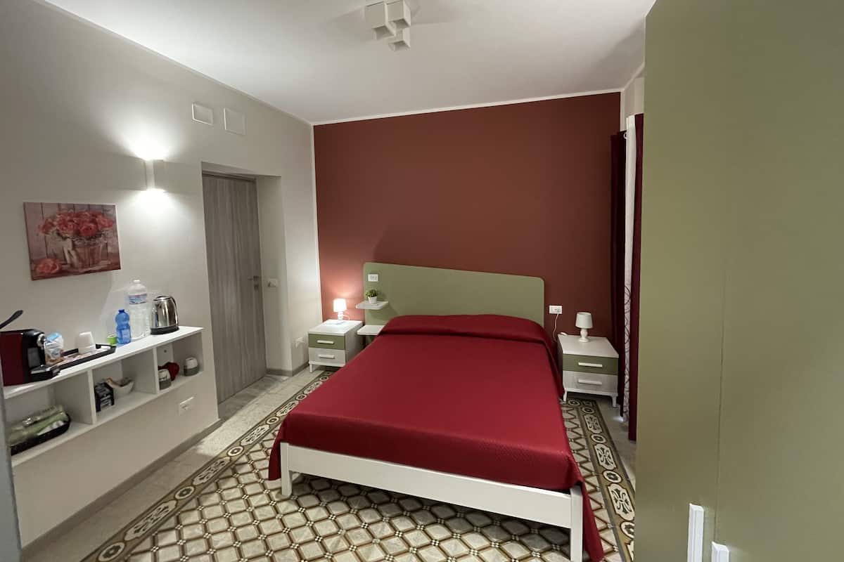 A bedroom with a bed, bedside tables, a nightstand, a shelf with toiletries, and a painting on the wall.