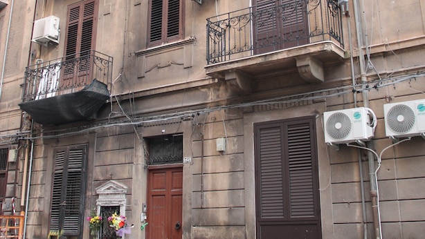 A red car parked in front of a building with multiple balconies, air conditioning units, and a wooden door. - Soffio di Relax (Palermo)
