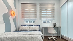 A bedroom with a bed, two windows with blinds, a desk with a chair, and a wall-mounted light. - Urban Designer Collection Lebuh Melaka (George Town)