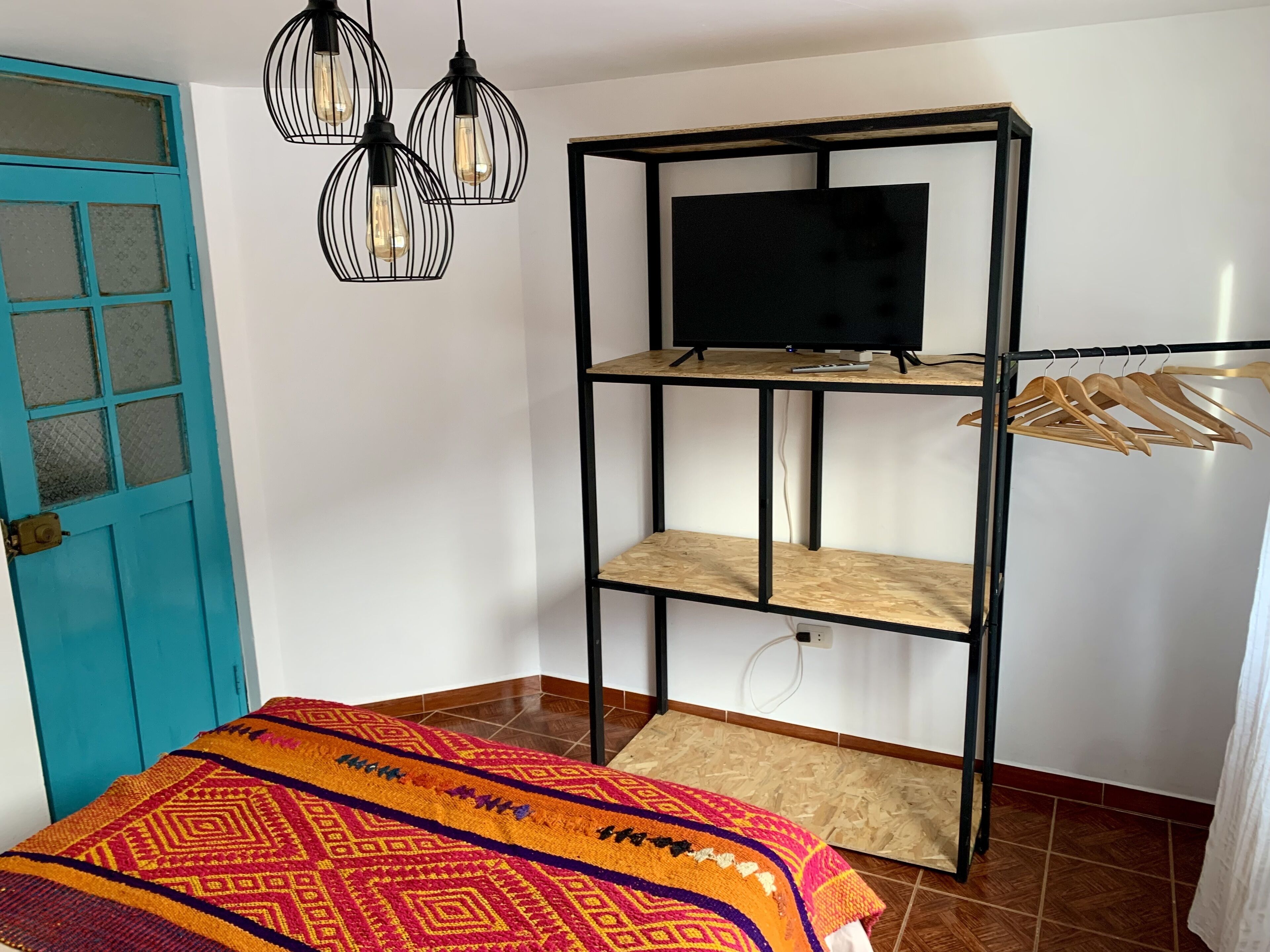Ein Zimmer mit blauer Tür, einem Bett mit gemustertem Überwurf, einem schwarzen Metall-TV-Schrank mit Fernseher und Holzkleiderhaken sowie Pendelleuchten.