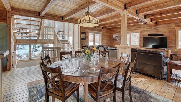 A log cabin interior with a dining area, a fireplace, a TV, and a staircase leading to a loft.