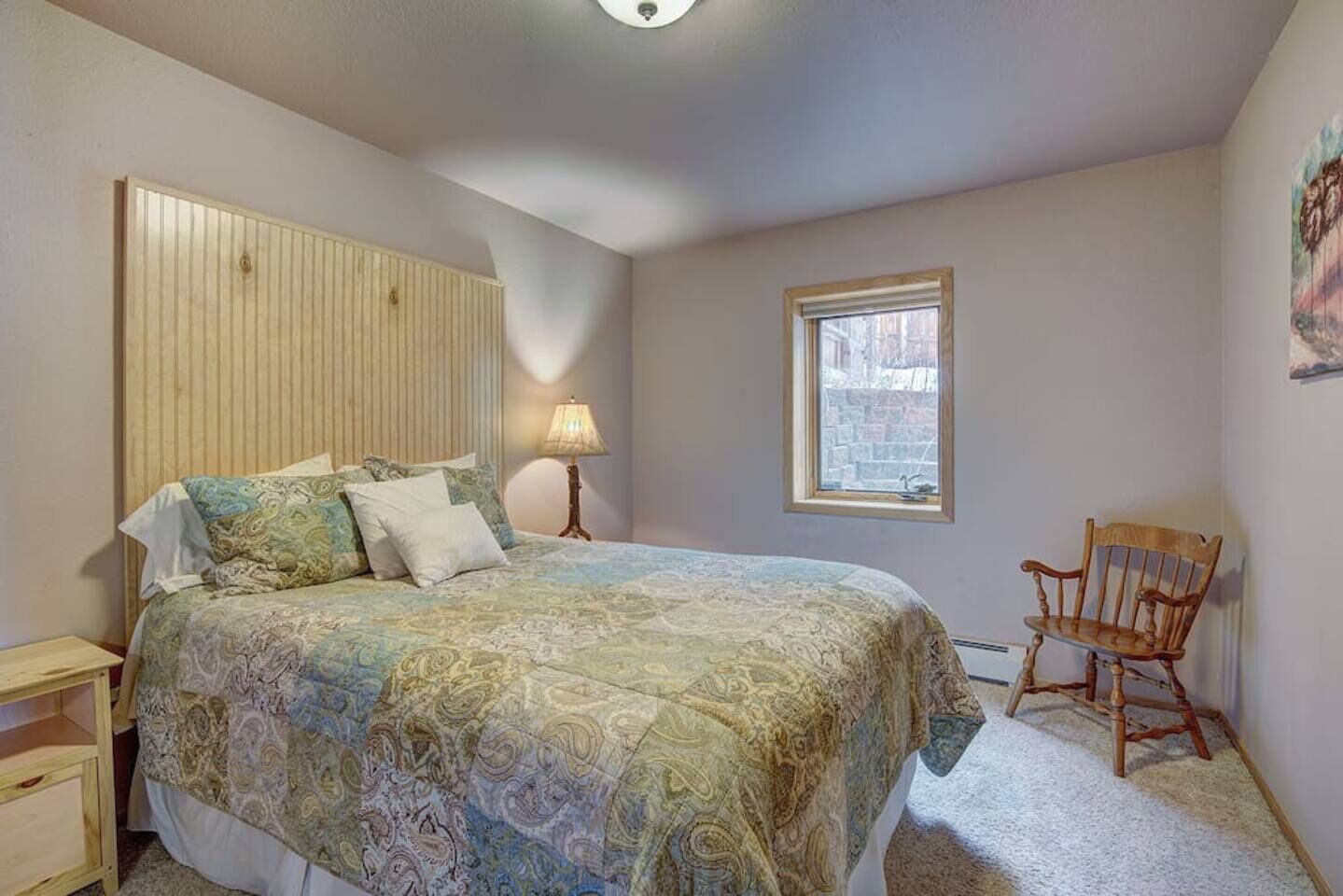 Une chambre à coucher comprenant un lit, une table de chevet, une lampe, une chaise, une fenêtre et une œuvre d’art accrochée au mur.
