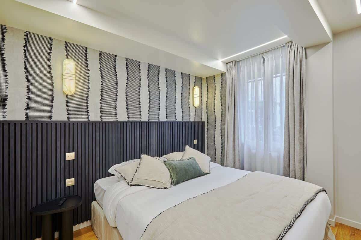 Un dormitorio moderno con cama, mesita de noche y una pared con rayas verticales e iluminación empotrada.