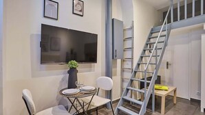 Ein modernes Wohnzimmer mit Flachbildfernseher, einem Esstisch mit Stühlen, einer Zimmerpflanze und einer Treppe.