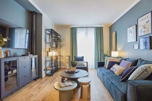 Una sala de estar moderna con un sofá, una mesa de centro y una mesita auxiliar. Se observa una estantería y un espejo en la pared.