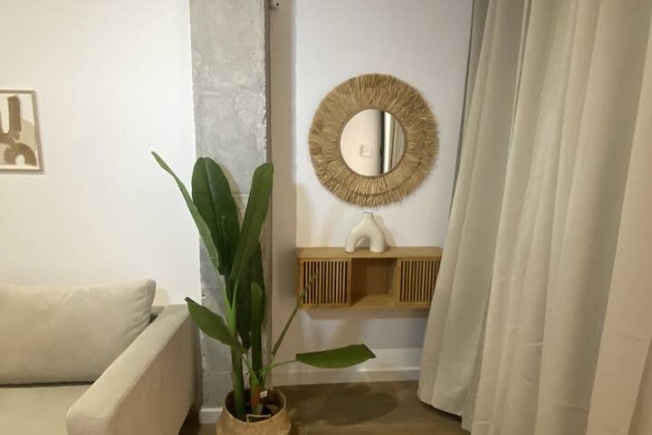 Et værelse med en beige sofa, en potteplante, et rundt spejl og en træhylde med en hvid vase.