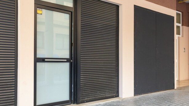 A building facade with closed roller shutters, a glass door with a handle, and a tiled floor.