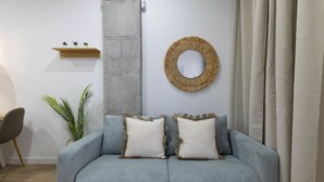 A modern living room with a grey sofa, two white pillows, a round woven mirror, and a small wooden shelf with plants.
