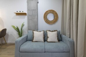 A modern living room with a grey sofa, two white pillows, a round woven mirror, and a small wooden shelf with plants.