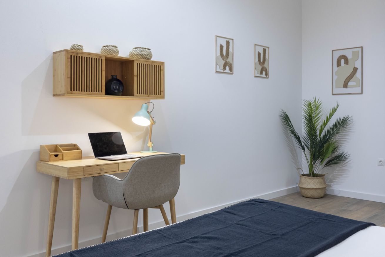 Een slaapkamer met een houten bureau, een laptop, een lamp, een bed met een blauwe deken, een potplant en wanddecoratie.