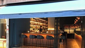 Un bar con mostrador y taburetes, una estantería llena de botellas, y una persona trabajando detrás de la barra.