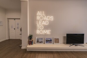 A modern interior with a neon sign stating "ALL ROADS LEAD TO ROME," a flat-screen TV, and framed pictures on a shelf. - Rhome Vintage Holidays San Pietro (Rome)