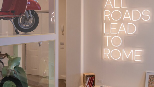 A modern interior with a neon sign stating "ALL ROADS LEAD TO ROME" on the wall, a red scooter, and a green scooter on display. - Rhome Vintage Holidays San Pietro (Rome)