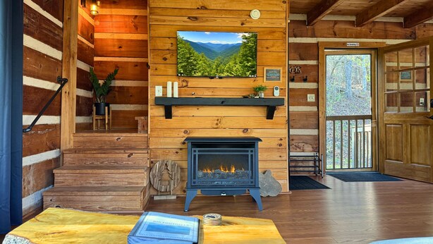 Interior acogedor de una cabaña con chimenea, paredes de madera, televisor montado y vistas al exterior.