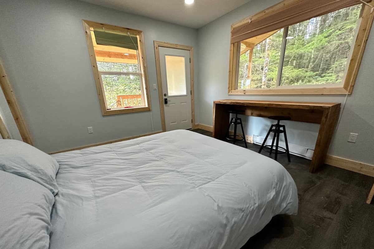 A bedroom with a large bed, a wooden desk with two stools, and a window with a forest view.