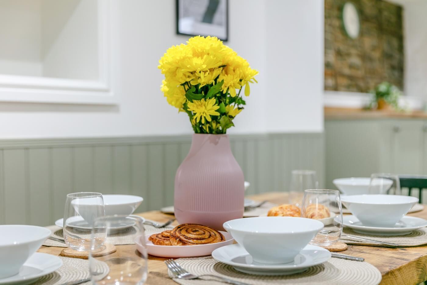 Une table à manger dressée avec de la vaisselle blanche, un vase rose contenant des fleurs jaunes et une assiette de petits pains à la cannelle.