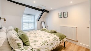 Une chambre à coucher comprenant un lit, un radiateur, une fenêtre avec des stores et un plafond avec des poutres apparentes.