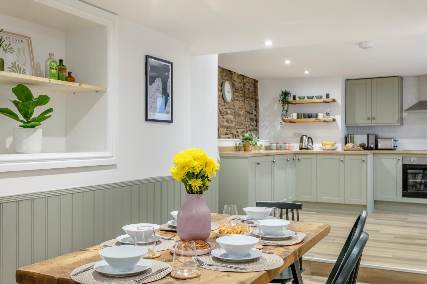 Comedor con mesa de madera dispuesta para cuatro personas, un florero con flores amarillas y una cocina al fondo.