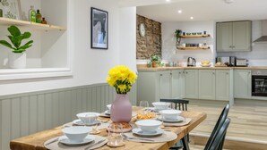 Un coin repas avec une table en bois dressée pour quatre personnes, un vase de fleurs jaunes et une cuisine en arrière-plan.