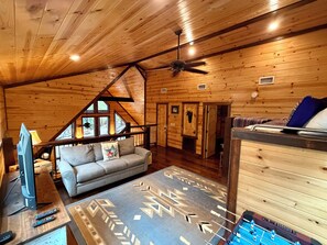 A cozy cabin interior with a loft bedroom, a living area with a sofa and a TV, and a game table. - Private Fishing Pond · Pedal Boat · Dogs · Hot Tub (Broken Bow)