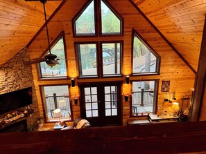 A cozy cabin interior with a stone fireplace, multiple windows, and a wooden ceiling.
