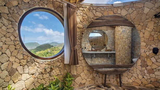 A stone-walled bathroom with a circular window, a mirror, and a shelf with toiletries. - Anima Eco Lodge (Kuta)
