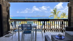 Un balcon avec une table et des chaises, donnant sur une plage aux eaux bleues et cristallines, bordée de palmiers.