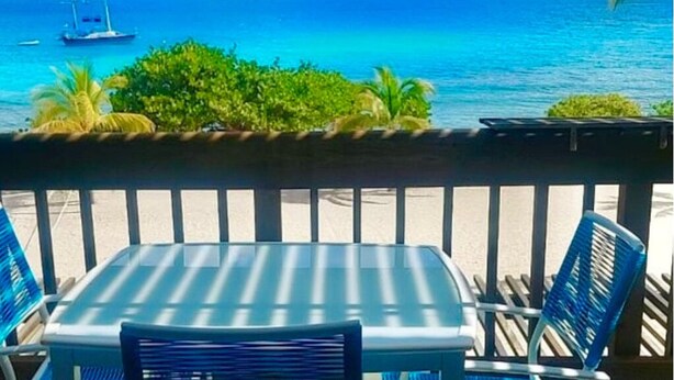 Un balcon avec une table et des chaises donnant sur une mer bleue et limpide, où l’on aperçoit un voilier.