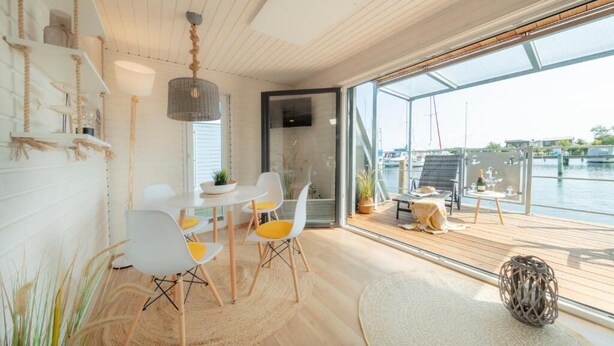 A modern interior with a dining area, a large window overlooking a body of water, and a wooden deck. - Hausboot Rán für 5 Pers. mit Großzügiger Dachterrasse in Kragenæs auf Lolland (Schleswig)