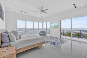 A modern bedroom with a large bed, a ceiling fan, and a view of the ocean through floor-to-ceiling windows. - Oceanfront Luxury Mansion, Dock, Pool&Spa, Kayaks (Marathon)