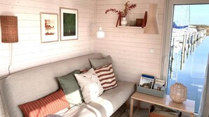 A cozy room with a wooden wall, a sofa with cushions, a small table, and a view of boats through a window.