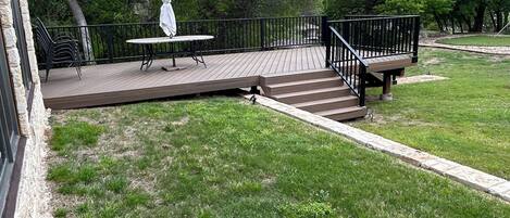 A wooden deck with a table and chairs, surrounded by a stone wall and a grassy lawn.