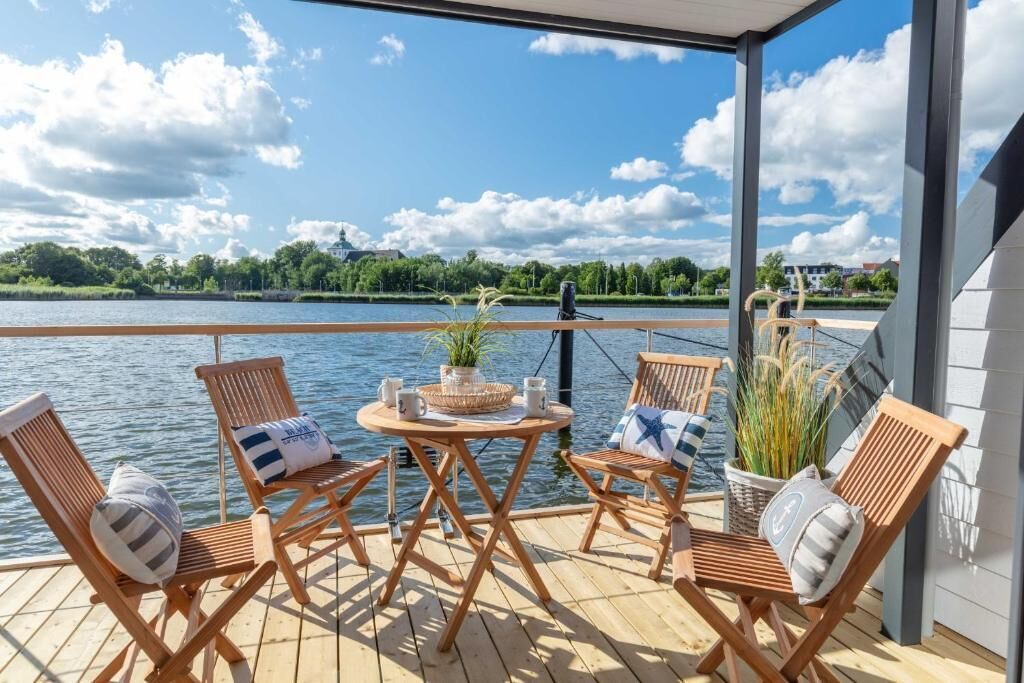 A wooden deck with four lounge chairs and a small table, overlooking a lake with a view of trees and buildings across the water.
