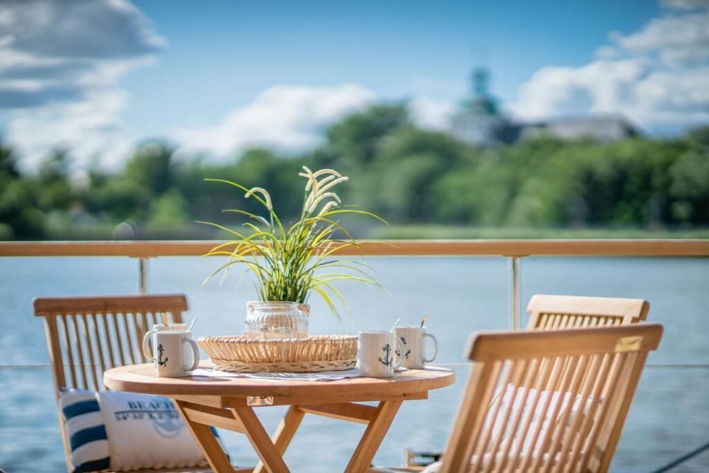 A wooden table with a potted plant, two mugs, and a woven basket on a balcony overlooking a body of water.