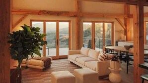 Un interior acogedor con sofá, reposapiés y zona de comedor, con vigas de madera y ventanas amplias con vistas a la montaña.