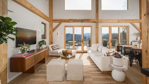 Amplia sala de estar con techos altos, vigas de madera, un televisor grande, un conjunto de sofás y un comedor con vistas a las montañas.
