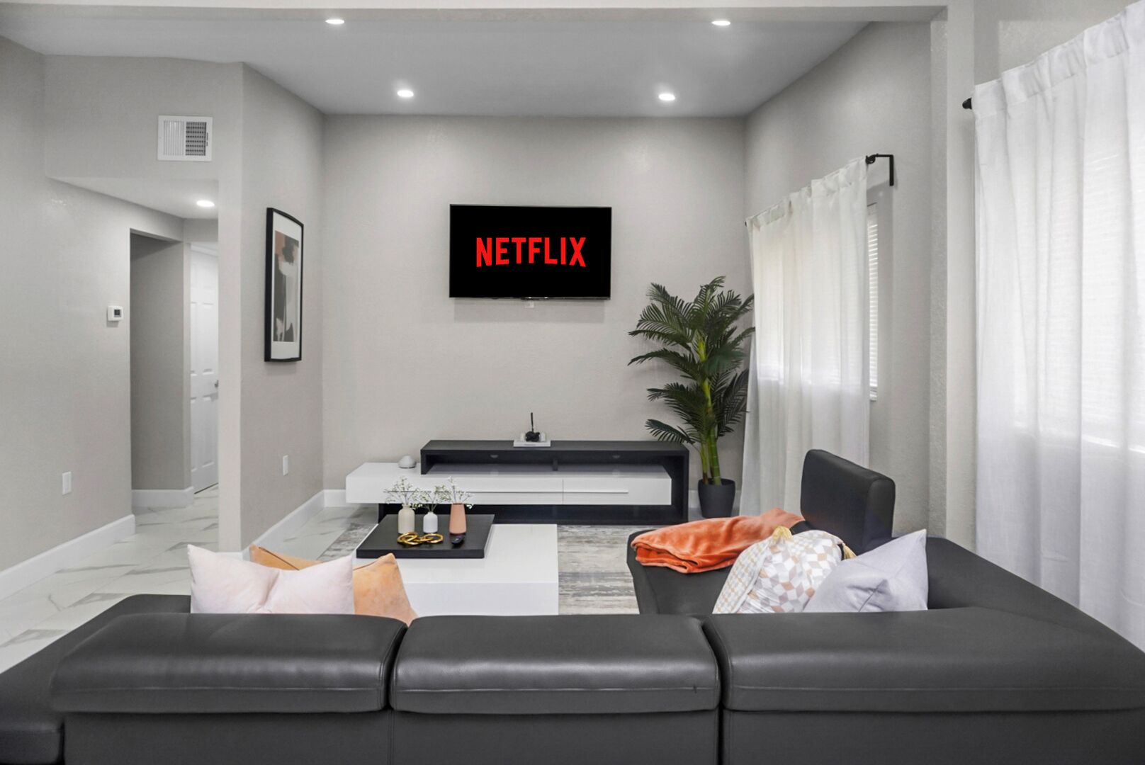 A modern living room with a black leather sofa, a coffee table, and a TV mounted on the wall displaying "NETFLIX".