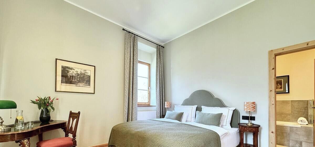 Une chambre à coucher avec un lit, un bureau avec une chaise, une fenêtre avec des rideaux et un tableau encadré au mur.