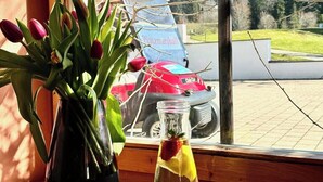 Blick aus dem Fenster auf einen Parkplatz mit einem roten Auto, einer Glasflasche mit Zitronenscheibe und einer Vase mit Tulpen.