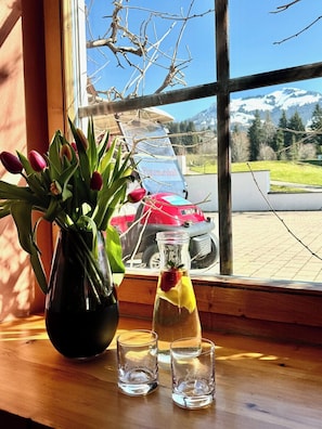 Udsigt fra et vindue over en parkeringsplads med en rød bil, en glasflaske med citronskive og en vase med tulipaner.