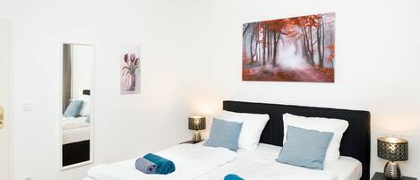 Ett modernt sovrum med en stor säng, två sängbord, en spegel och en tavla med skogsmotiv.