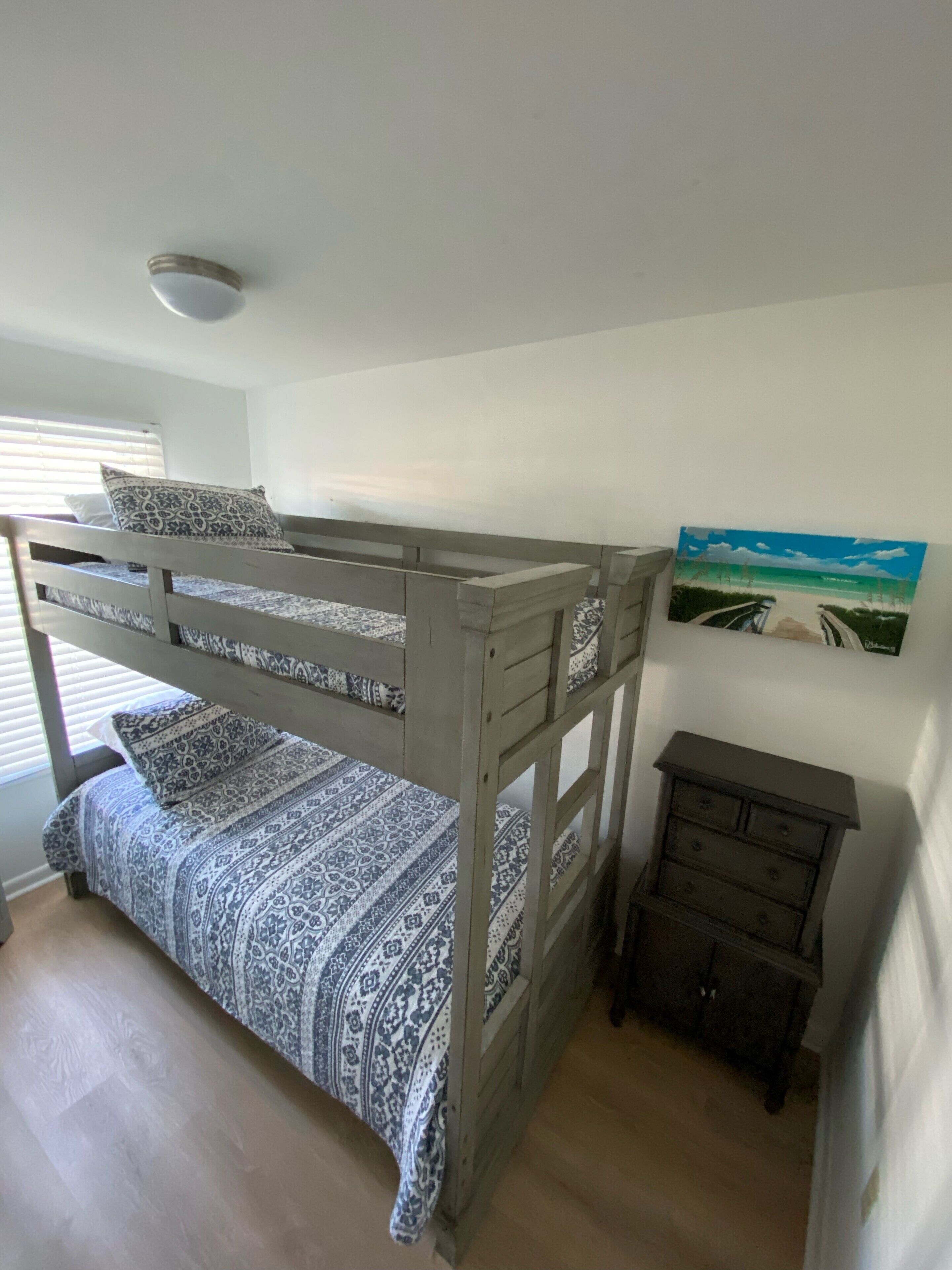 A bunk bed with patterned bedding, a small dresser, and a painting of a beach scene.