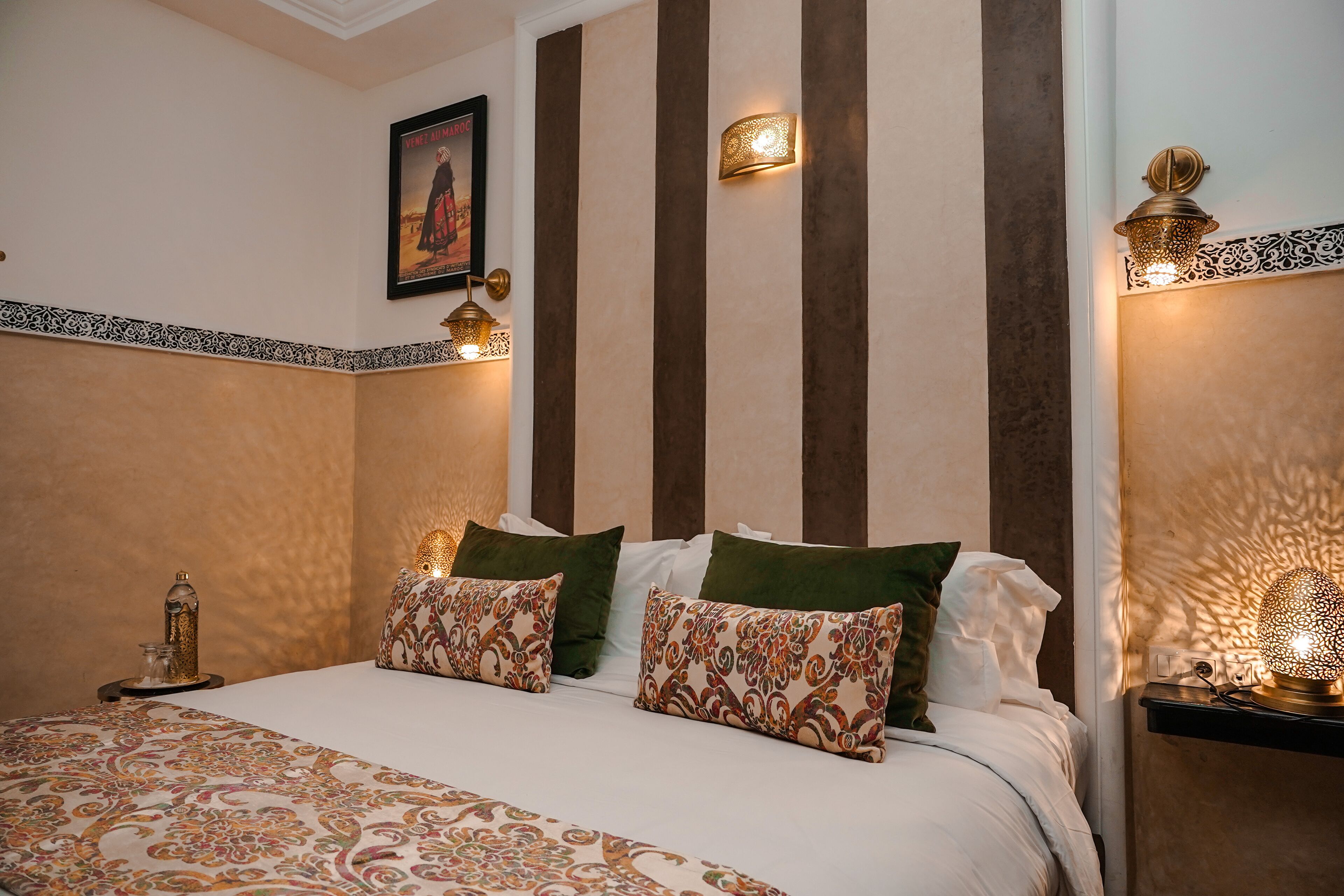 A hotel room with a bed, decorative pillows, wall-mounted lamps, and a framed picture on the wall.